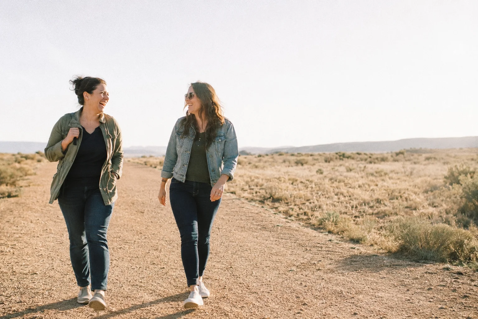 Two friends walking together on an open path in bright daylight, one laughing